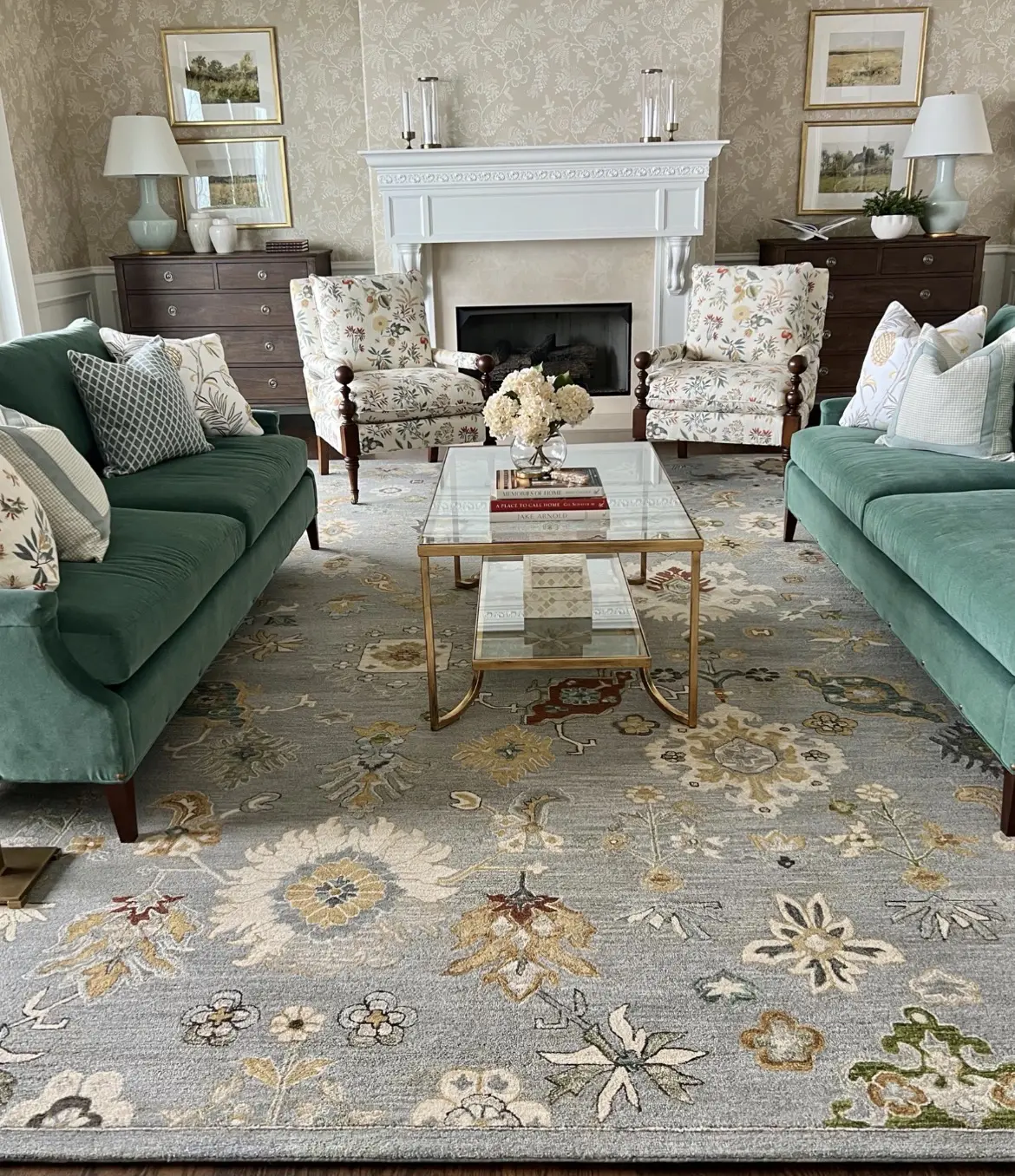Two teal sofas face a glass coffee table in a classic living room with floral armchairs, a white fireplace, and wallpapered walls.