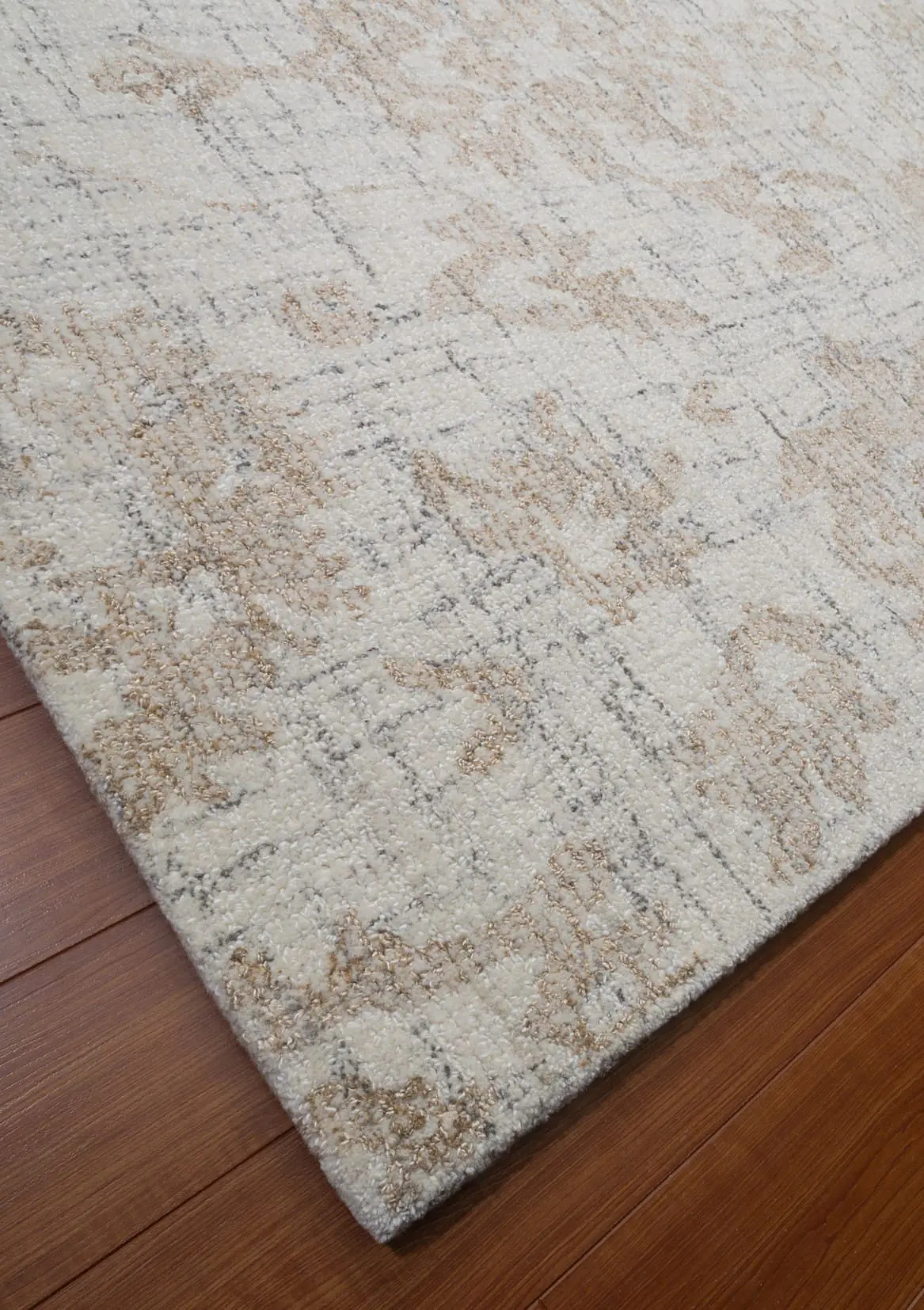 Corner of a light beige area rug with gray and tan pattern on a wooden floor.
