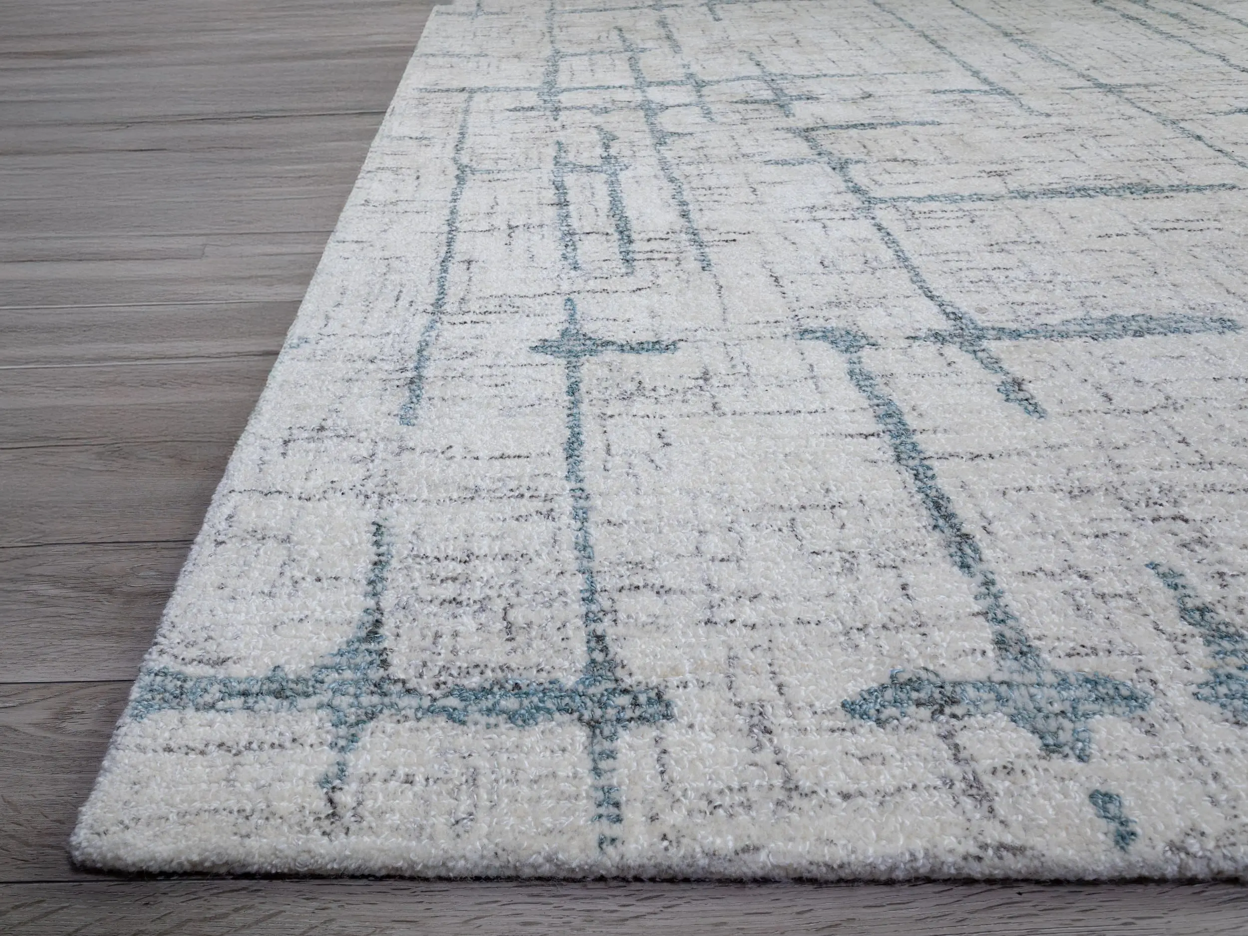 Light cream rug with a blue rectangular grid pattern laid over a wooden floor, showing the rug edge.