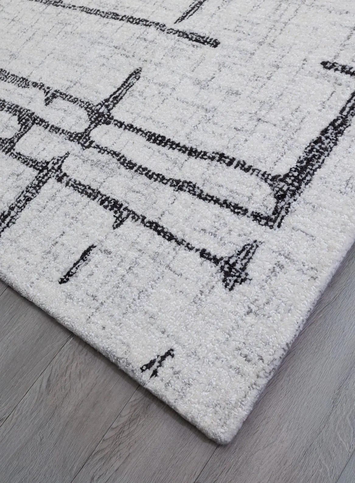 Close-up of a light gray plush rug with a black geometric border pattern on a gray wooden floor.